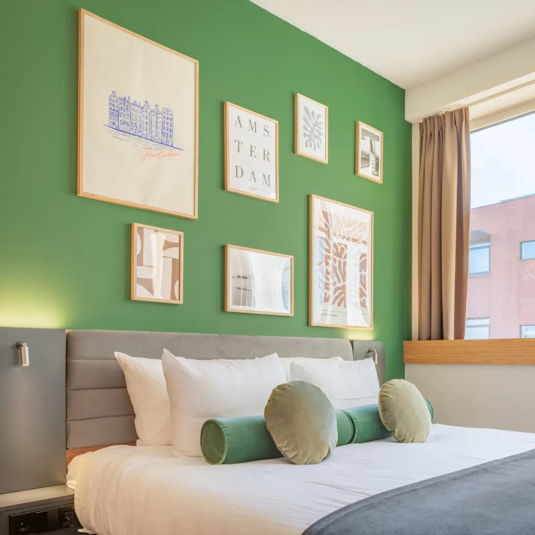 Moderne hotelkamer met groene accentmuur, grijs gestoffeerd hoofdeinde, wit beddengoed en Amsterdams ingelijst kunstwerk